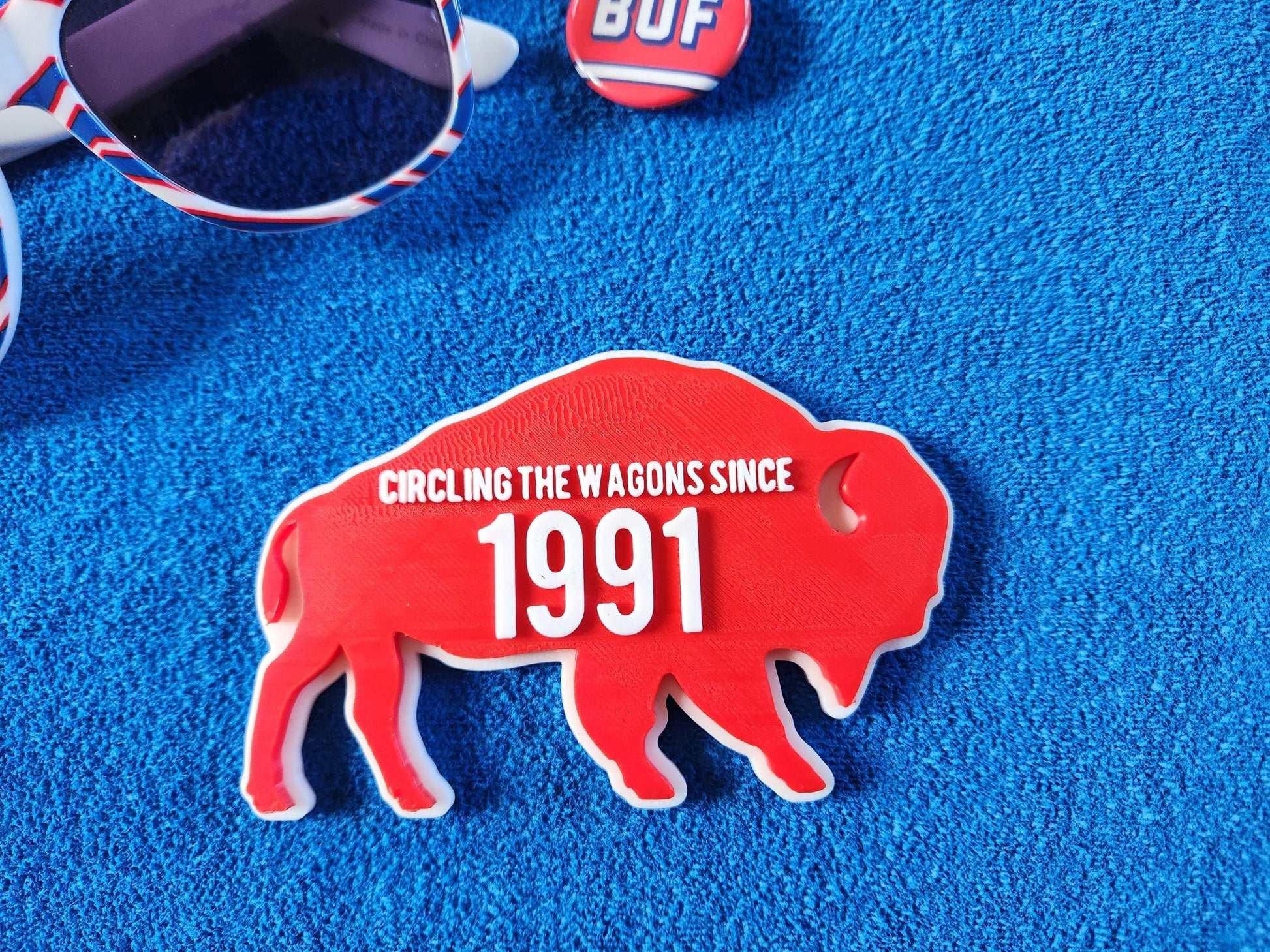 A red magnet for Buffalo Bills fans that says Circling the Wagons since 1991 that can be customized with any year. It is shown on a blue background next to some accessories like keys and a pin.