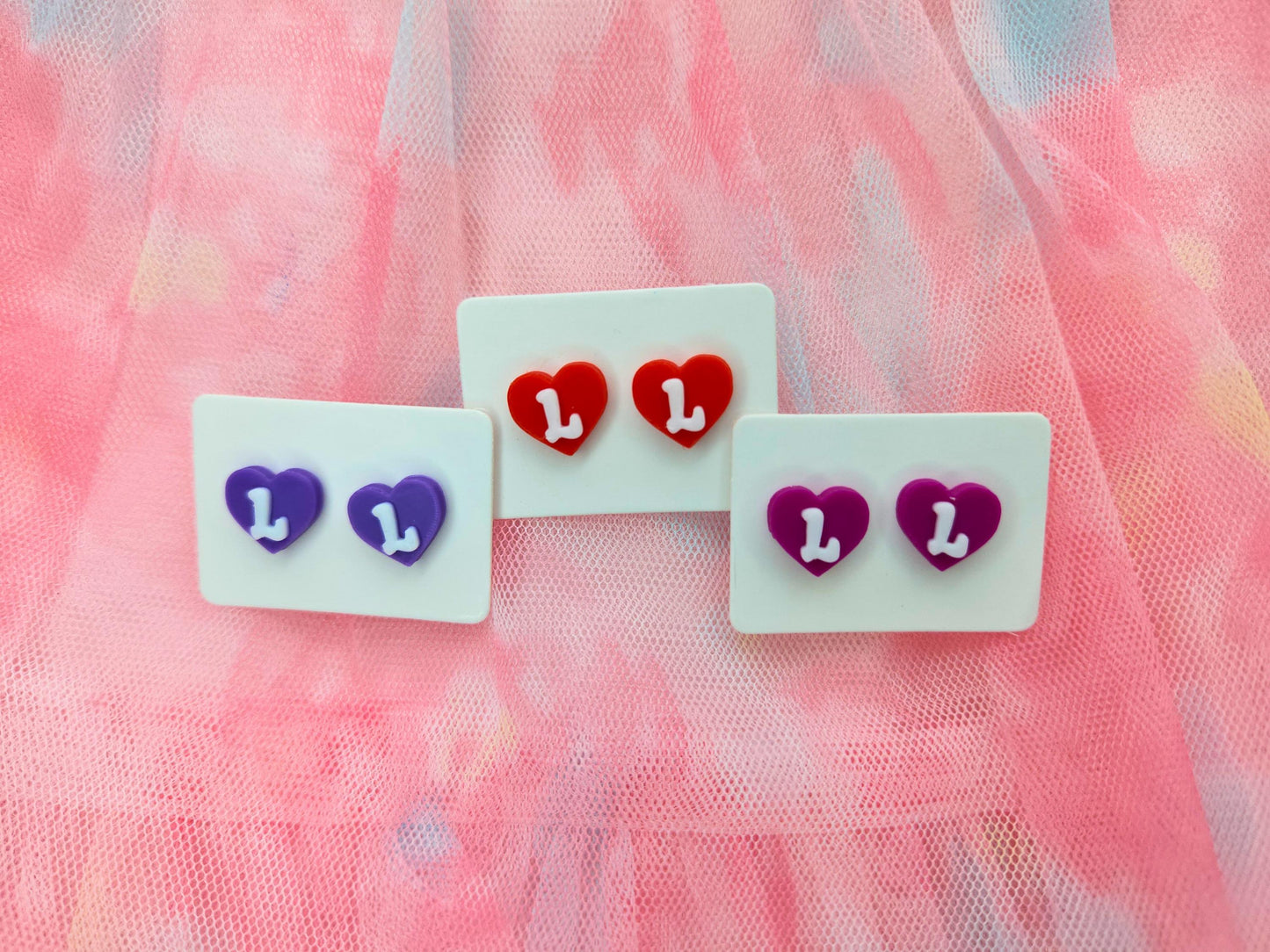 Three pairs of heart-shaped stud earrings with the letter "L" displayed on a soft pink tulle background. Earrings are red, purple, and magenta.