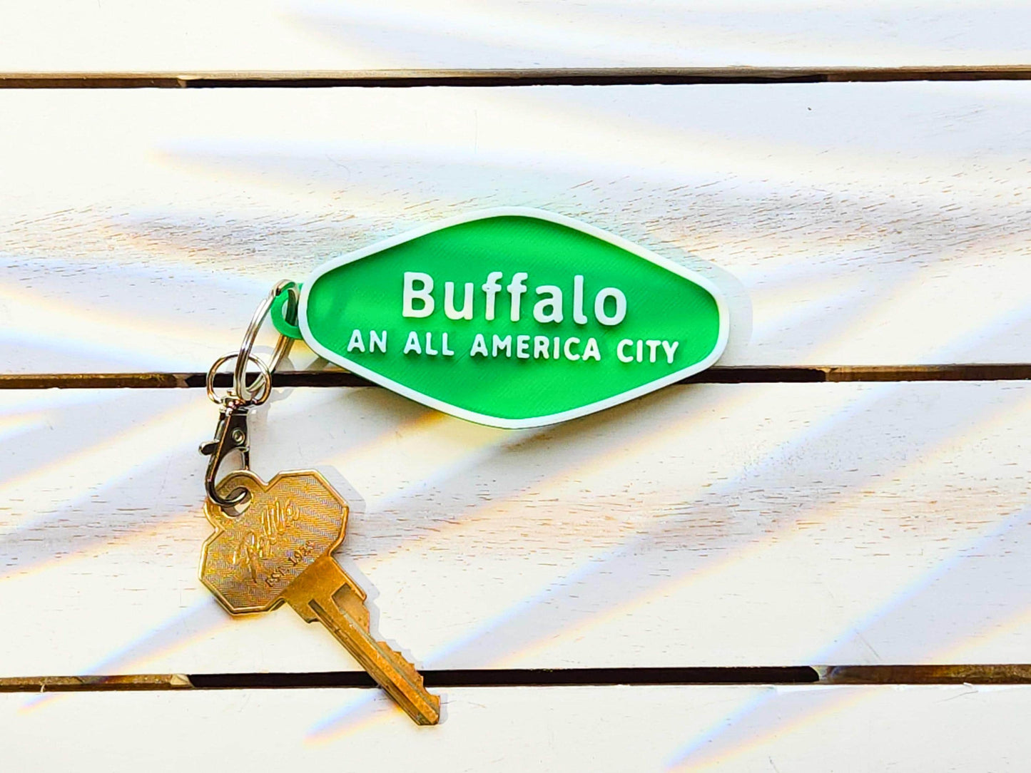 A green 3D-printed diamond shaped Buffalo keychain with white raised text, on a wooden background. The keychain says Buffalo An All America City and is shown with a key for scale.