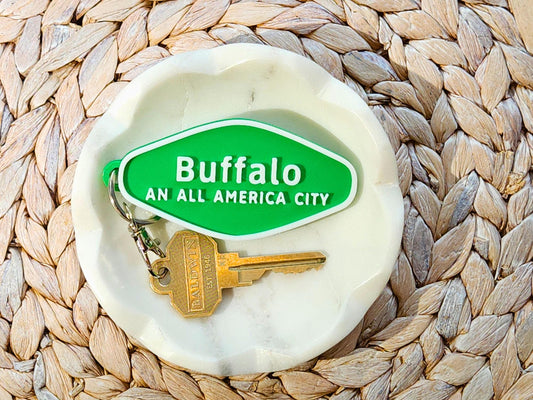 A green 3D-printed diamond shaped Buffalo keychain with white raised text, in a white dish on a wooden background. The keychain says Buffalo An All America City and is shown with a key for scale.