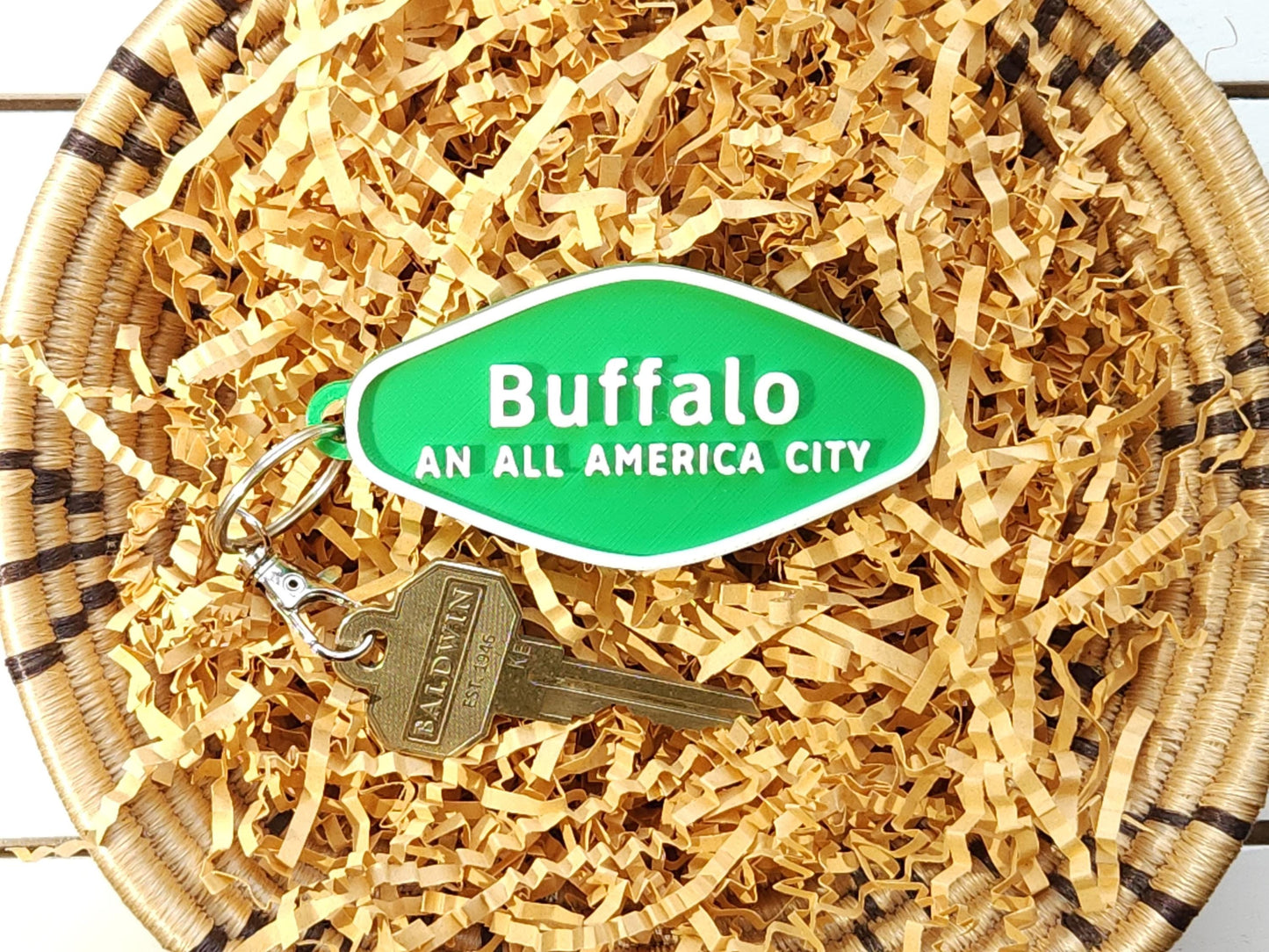A green 3D-printed diamond shaped Buffalo keychain with white raised text, in a basket on a wooden background. The keychain says Buffalo An All America City and is shown with a key for scale.
