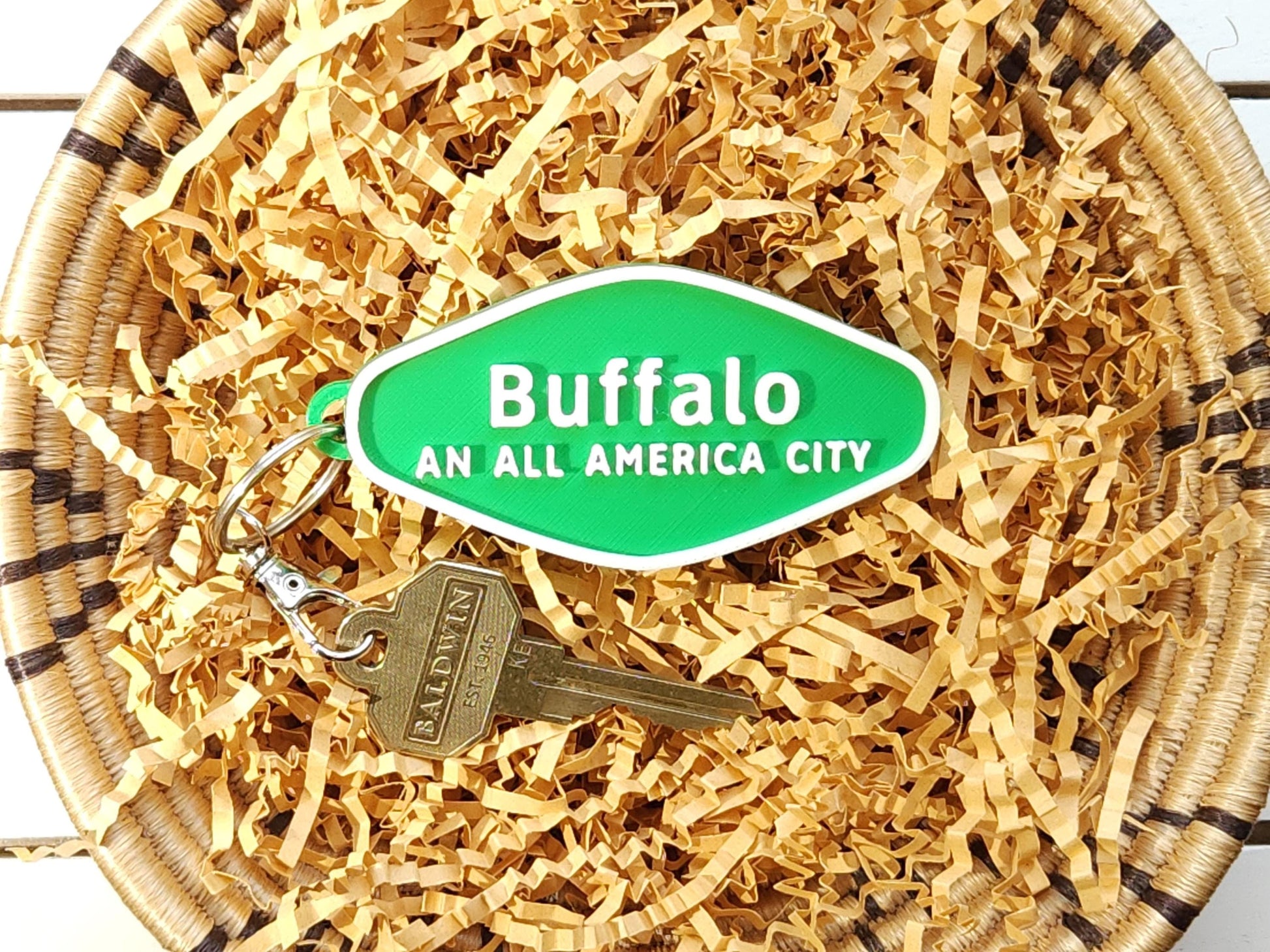 A green 3D-printed diamond shaped Buffalo keychain with white raised text, in a basket on a wooden background. The keychain says Buffalo An All America City and is shown with a key for scale.