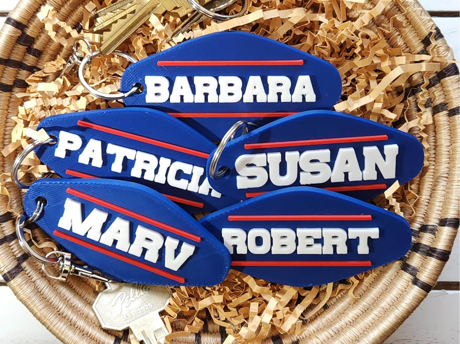 A set of 3 personalized keychains, done in a retro Buffalo Bills style that says the names PATRICIA, BARBARA, MARV, ROBERT and SUSAN in all caps with a red line above and below it on a blue background. The keychains are in a basket.
