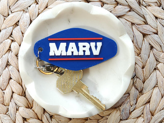 A personalized keychain, done in a retro Buffalo Bills style that says the name MARV in all caps with a red line above and below it on a blue background. The keychain is in a white dish on a woven background.