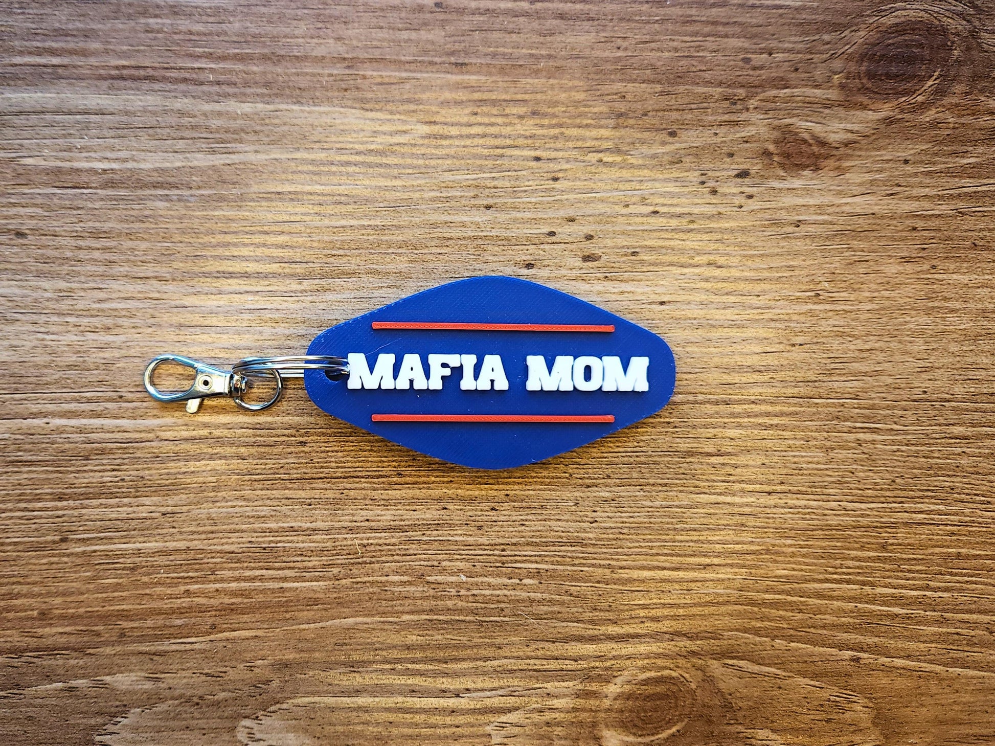 A personalized keychain, done in a retro Buffalo Bills style that says the names MAFIA MOM in all caps with a red line above and below it on a blue background. The keychain is on a wooden background.