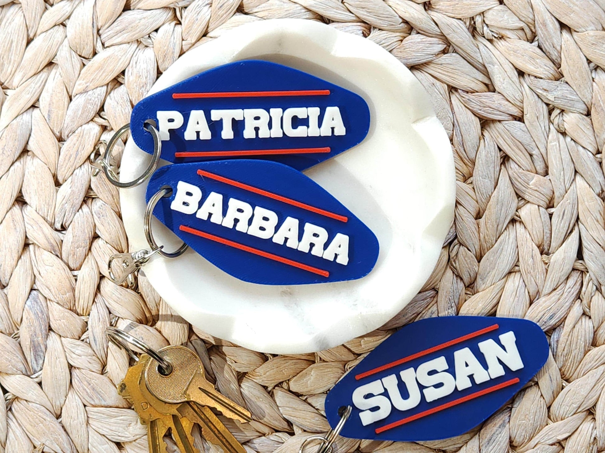 A set of 3 personalized keychains, done in a retro Buffalo Bills style that says the names PATRICIA, BARBARA and SUSAN in all caps with a red line above and below it on a blue background. The keychains are in a white dish on a woven background.