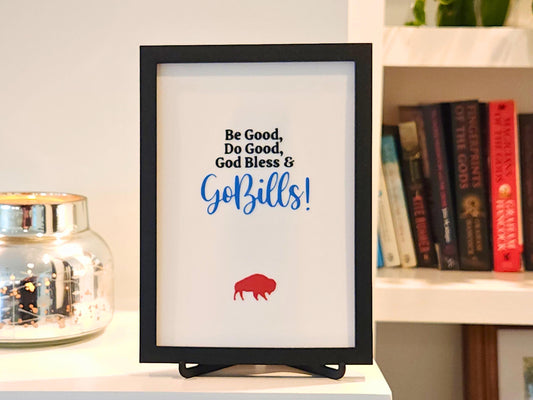 A rectangular 3D printed sign in a matte black frame, featuring the quote “Be Good, Do Good, God Bless & Go Bills!” in black and Bills blue text, with a bold red buffalo icon underneath. The sign is displayed on a coordinating black 3D printed easel stand. In the background is a silver mercury glass candle holder and a built-in bookshelf with neatly arranged books and plants, adding a cozy home vibe.