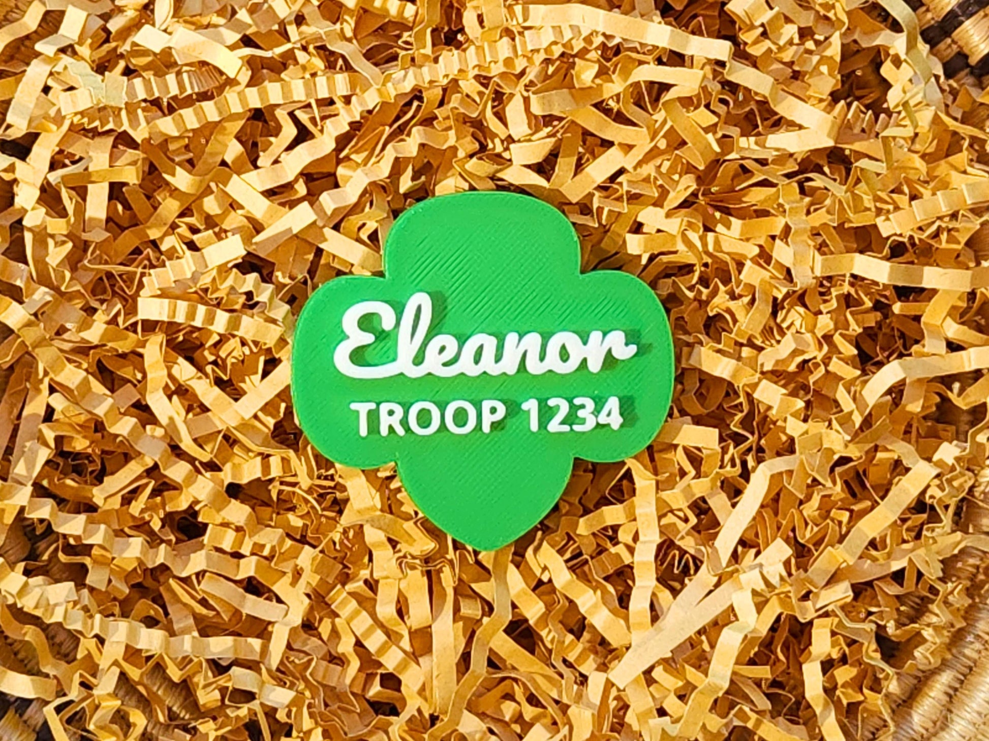 A personalized green Girl Scout-shaped magnet displayed in basket of decorative grass. It features the name 'Eleanor' in a white script font and 'TROOP 1234' underneath in bold capital letters