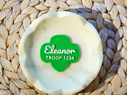 Personalized Girl Scout pin in green trefoil shape with name "Eleanor" and Troop 1234 in white text, displayed in a marble dish—custom gift for Girl Scouts.
