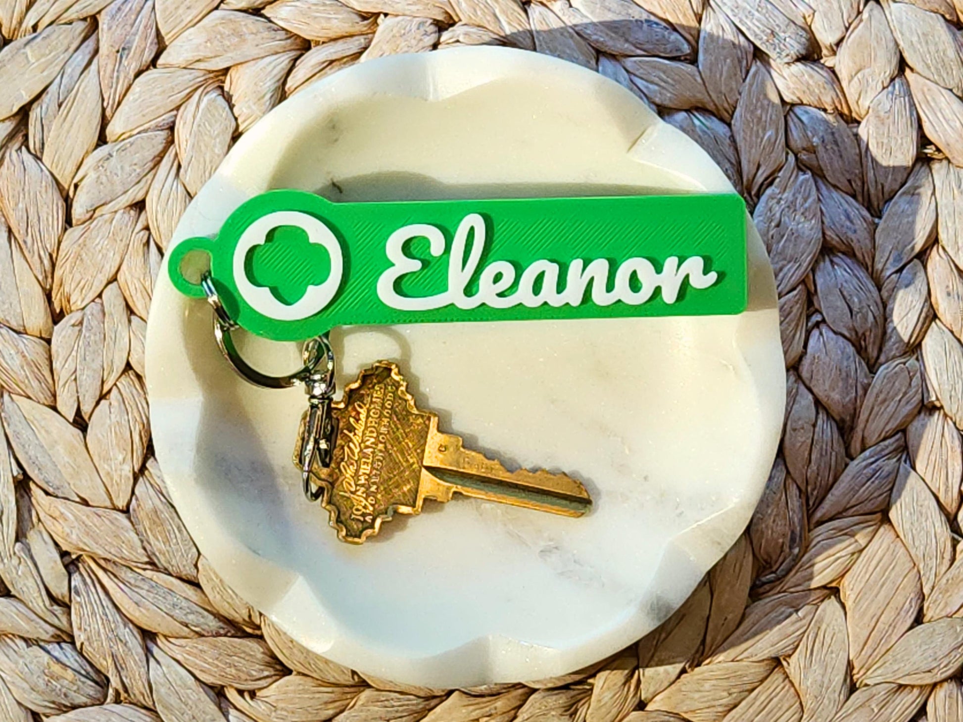 Personalized Girl Scout keychain with name “Eleanor” in white script, printed on a green rectangular tag featuring the Girl Scout trefoil logo, shown with a house key on a white marble dish—great for backpacks, troop leaders, and scout gifts.