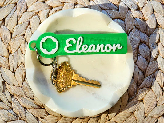 Personalized Girl Scout keychain with name “Eleanor” in white script, printed on a green rectangular tag featuring the Girl Scout trefoil logo, shown with a house key on a white marble dish—great for backpacks, troop leaders, and scout gifts.