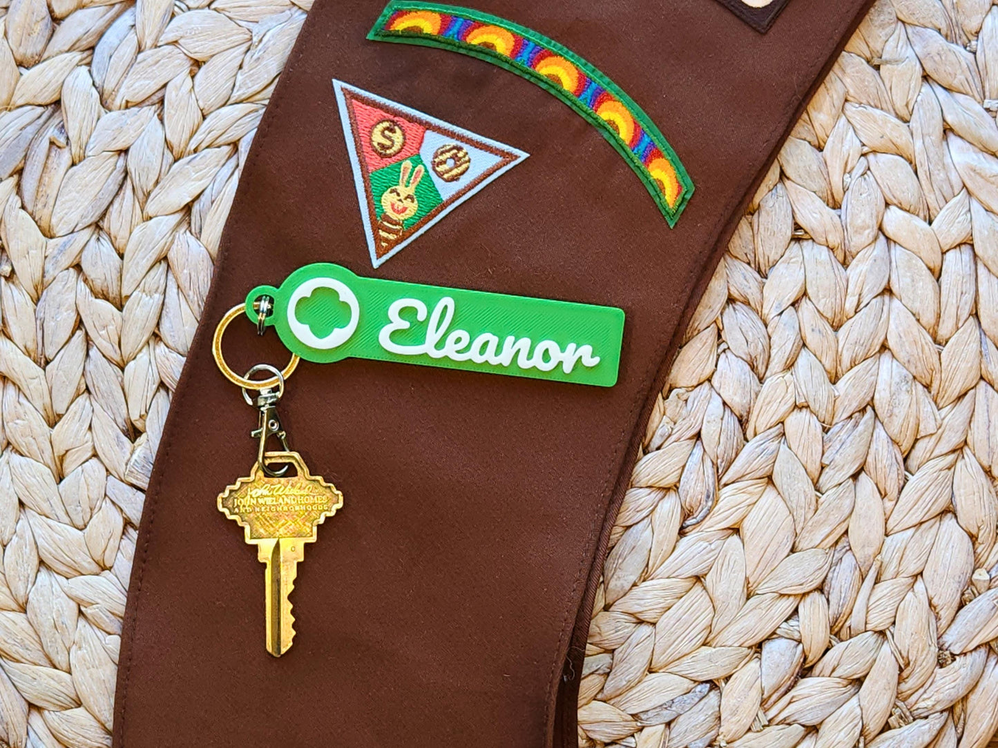Custom Girl Scout name keychain in green with white trefoil emblem and personalized “Eleanor” script text, displayed on a Brownie sash with embroidered patches—ideal for Girl Scout swaps, cookie season gifts, or personalized troop gear.