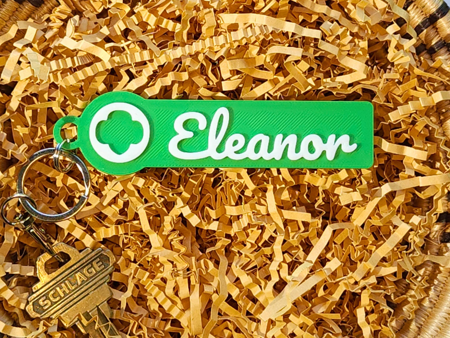 Green personalized Girl Scout keychain nestled in basket filler, showing white script name “Eleanor” with trefoil logo—thoughtful end-of-year Girl Scout gift, bridging ceremony favor, or troop leader appreciation idea.