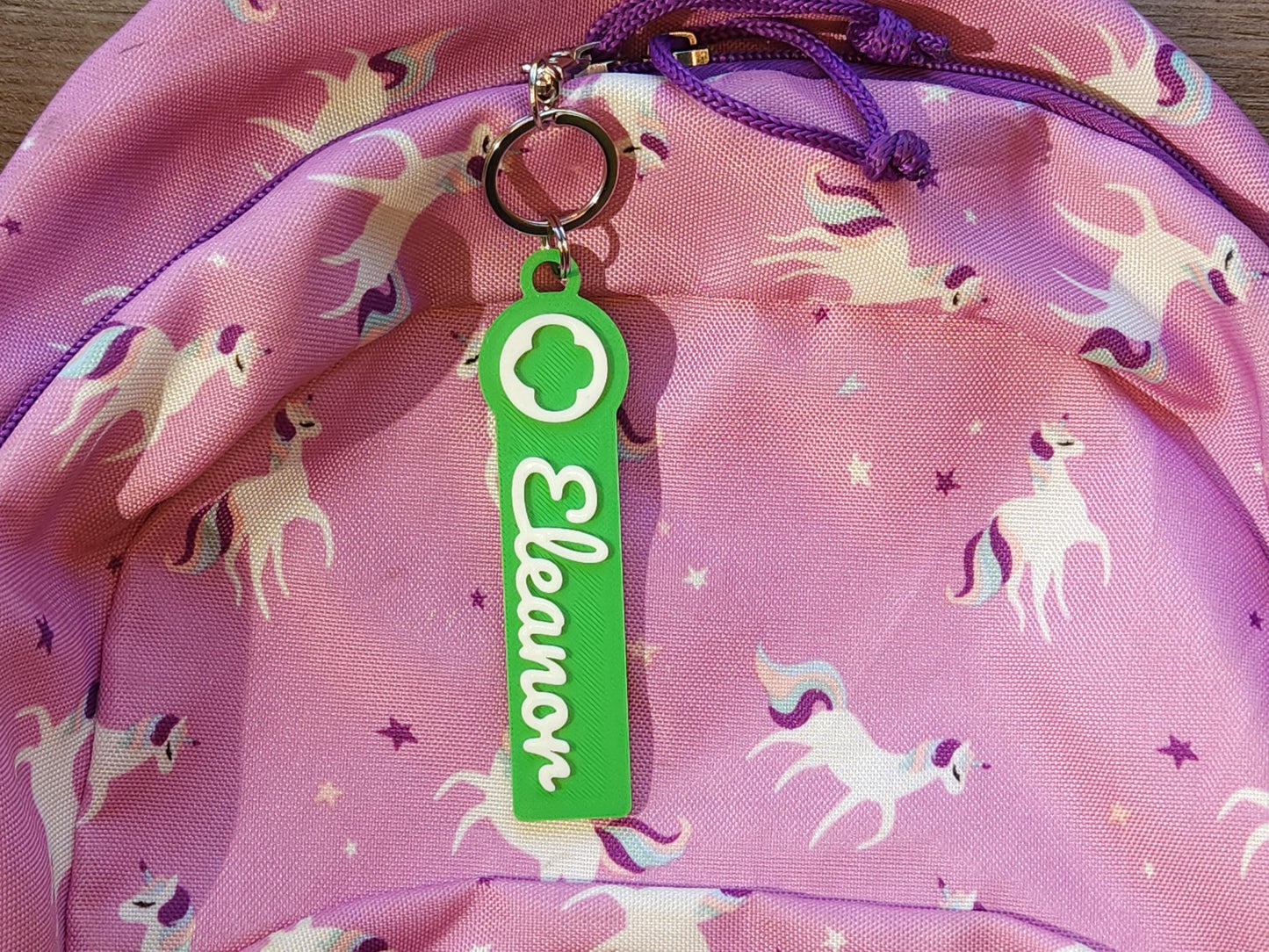 Girl Scout personalized keychain for girls, featuring green name tag with white “Eleanor” script and Girl Scout logo, clipped to a pink unicorn backpack—fun and functional for school bags, jackets, or scout totes.