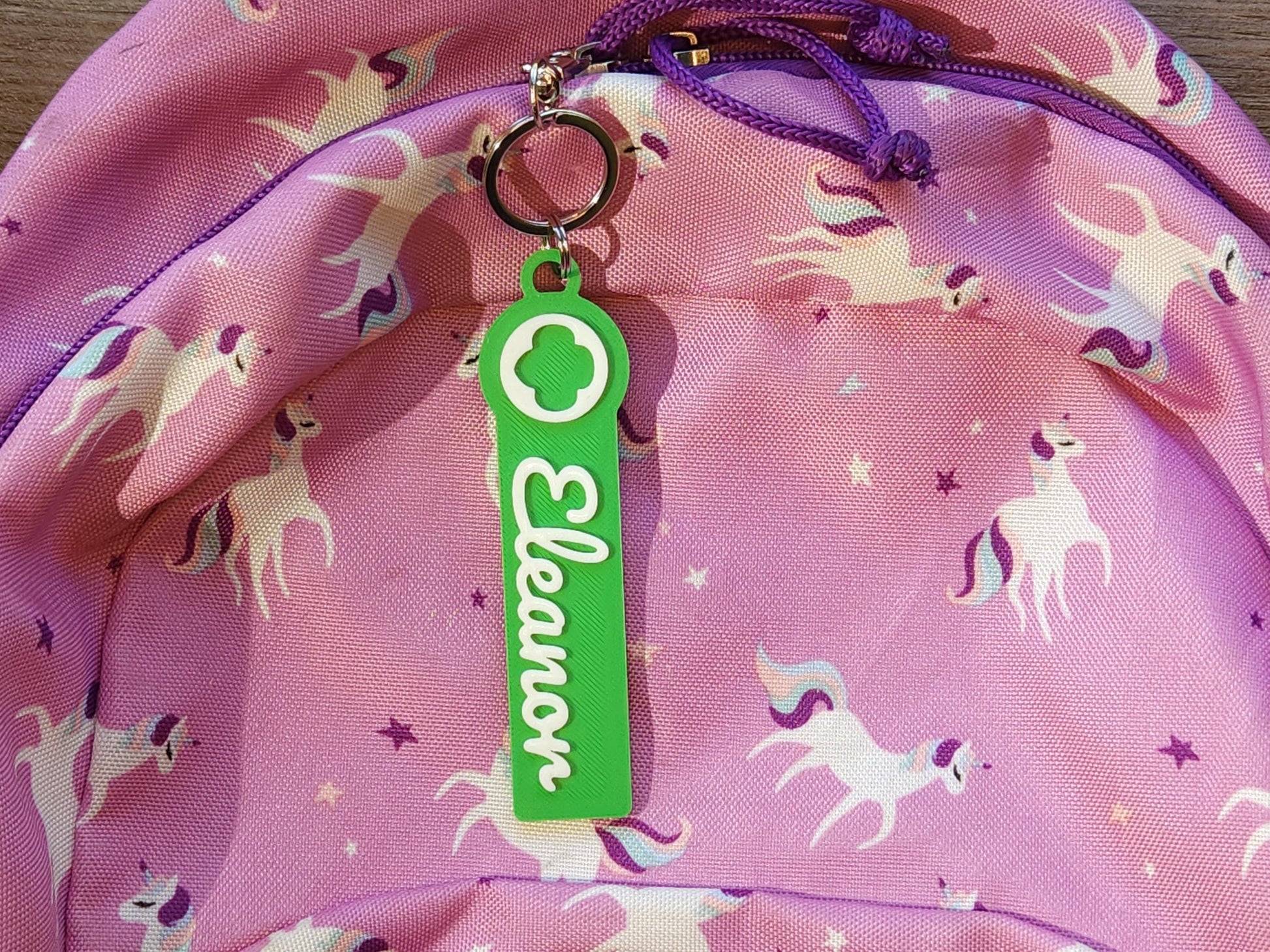 Girl Scout personalized keychain for girls, featuring green name tag with white “Eleanor” script and Girl Scout logo, clipped to a pink unicorn backpack—fun and functional for school bags, jackets, or scout totes.