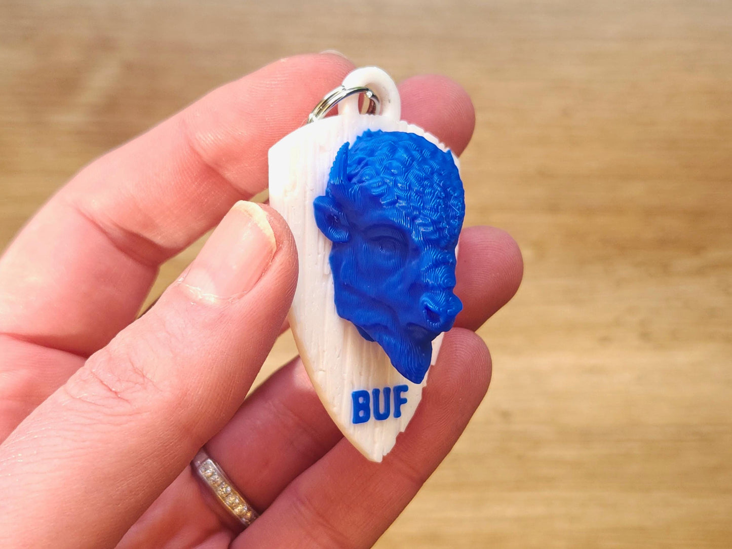 Close-up of a blue 3D printed buffalo head on a faux wood white shield keychain, held in hand. "BUF" is printed below in blue.