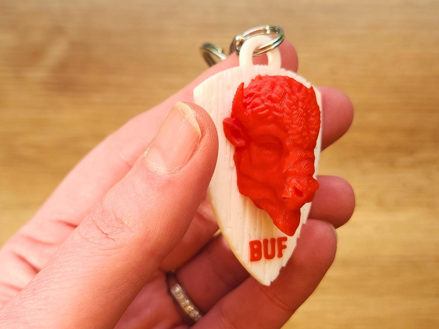 Close-up of a red 3D printed buffalo head on a faux wood white shield keychain, held in hand. The letters "BUF" are printed below the buffalo in red.