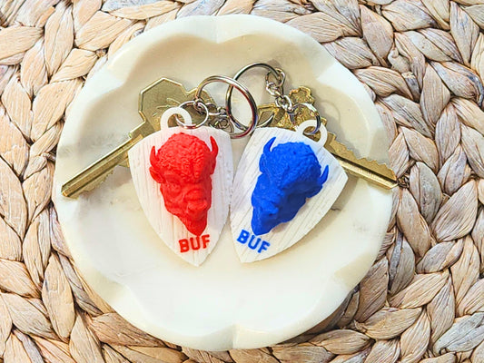 Flat lay of two buffalo keychains (one red, one blue) on a small dish, attached to metal key rings with keys.