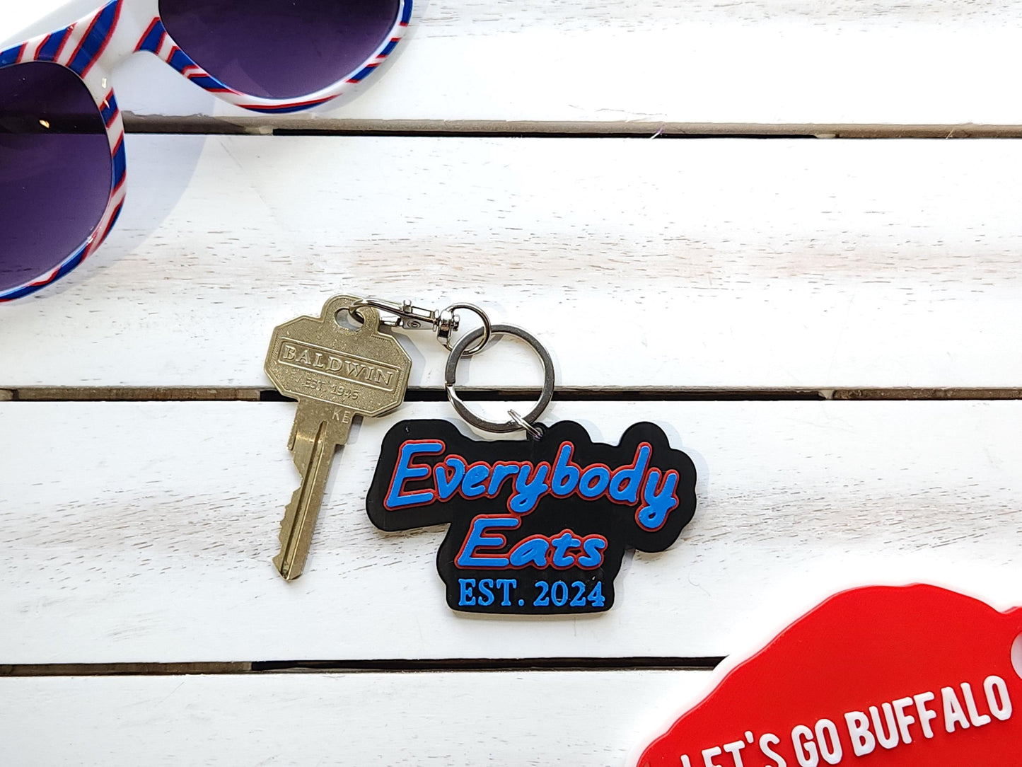Buffalo Bills "Everybody Eats" Est. 2024 keychain next to red, white, and blue sunglasses on white wooden surface – tailgate and sports fan accessory