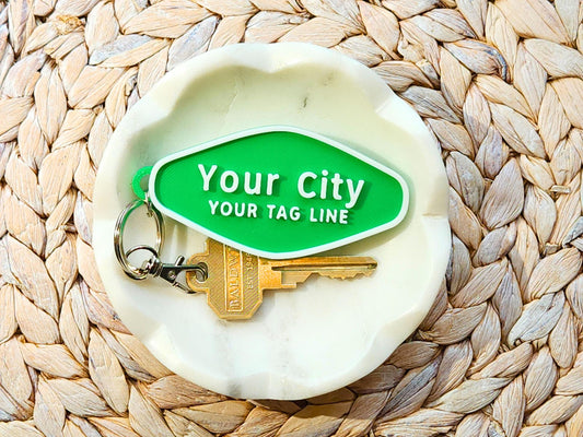 Close-up of a single personalized keychain that reads “Your City, Your Tag Line” with keyring and brass key on a white dish and woven placemat.