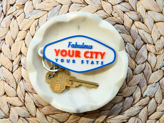 Top-down view of custom 3D printed YOUR CITY keychain in marble catchall tray on woven placemat background