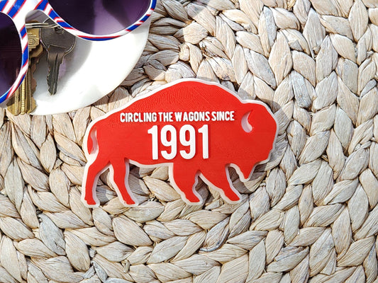 A red magnet for Buffalo Bills fans that says Circling the Wagons since 1991 that can be customized with any year. It is shown on a woven background next to some accessories like keys and sunglasses.
