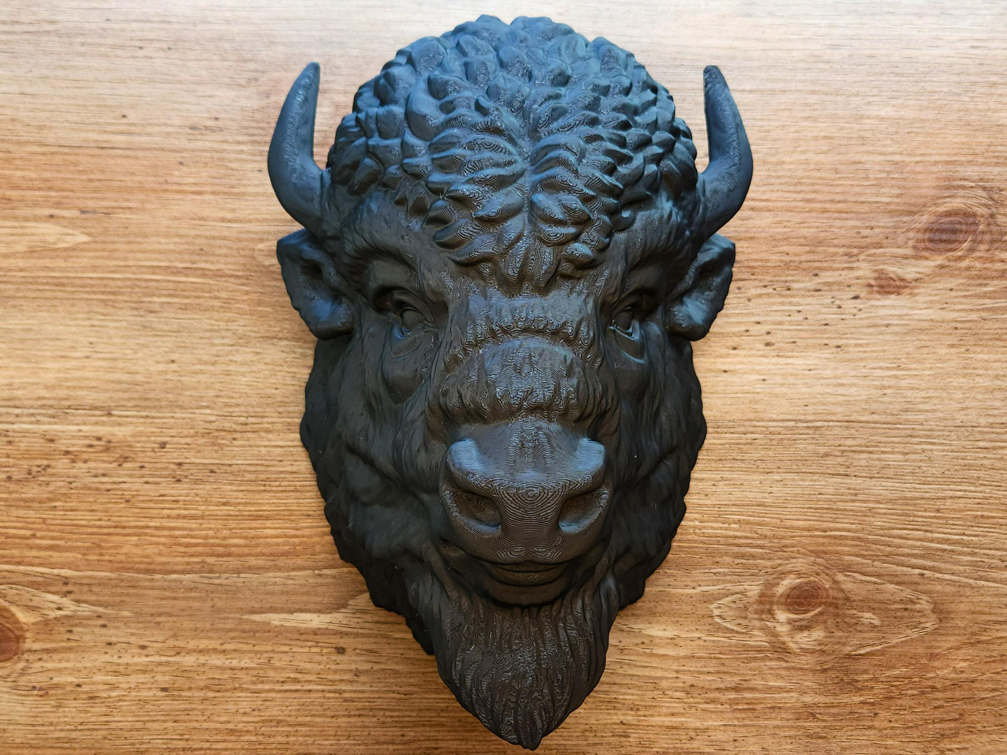 Front view of a detailed 3D printed black buffalo head wall sculpture with textured fur, horns, and lifelike facial features, displayed on a wooden surface.