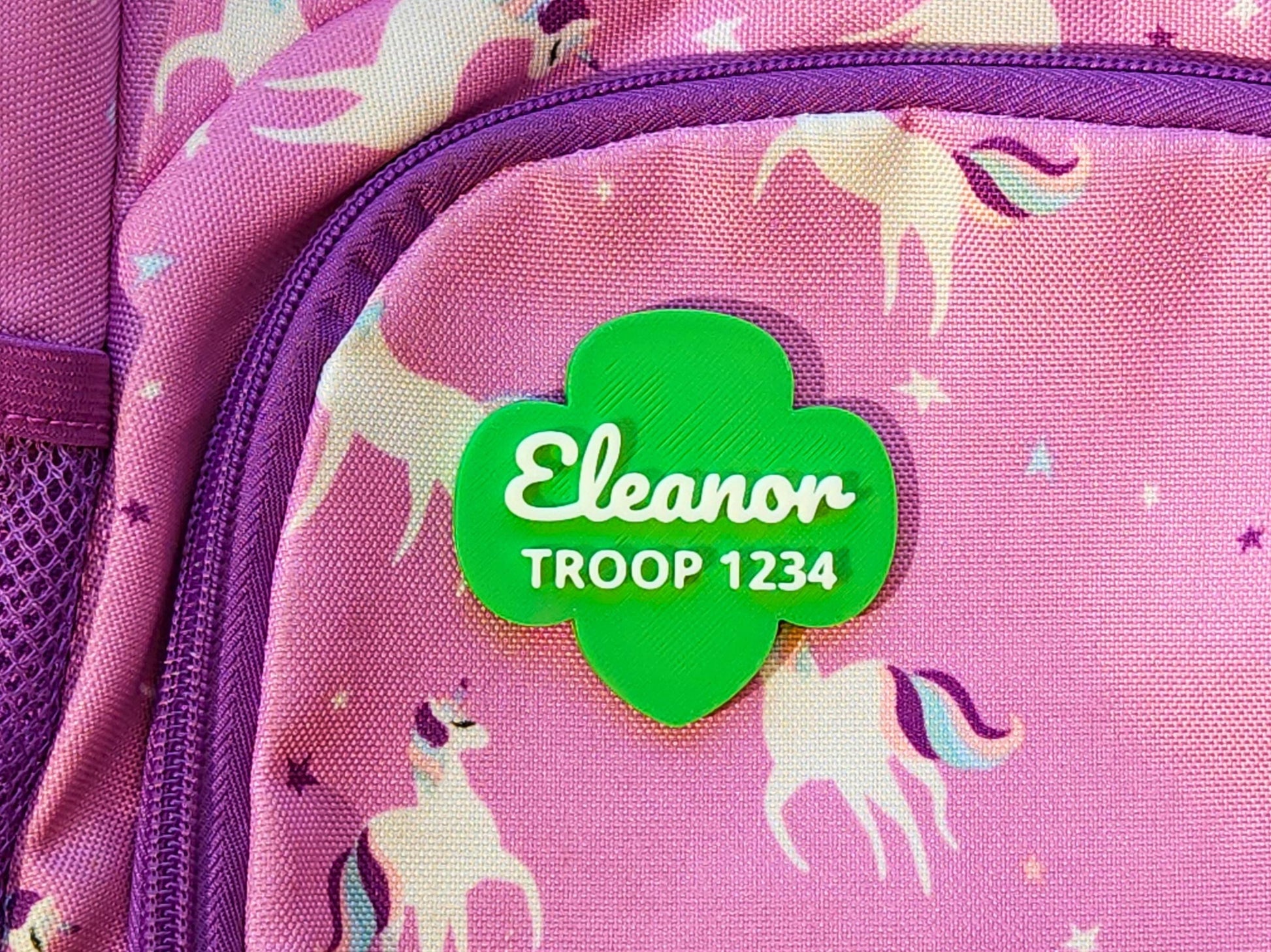 Custom green Girl Scout pin with white lettering for name and troop number, designed in the iconic trefoil shape and displayed on a backpack—great for end-of-year troop gifts.