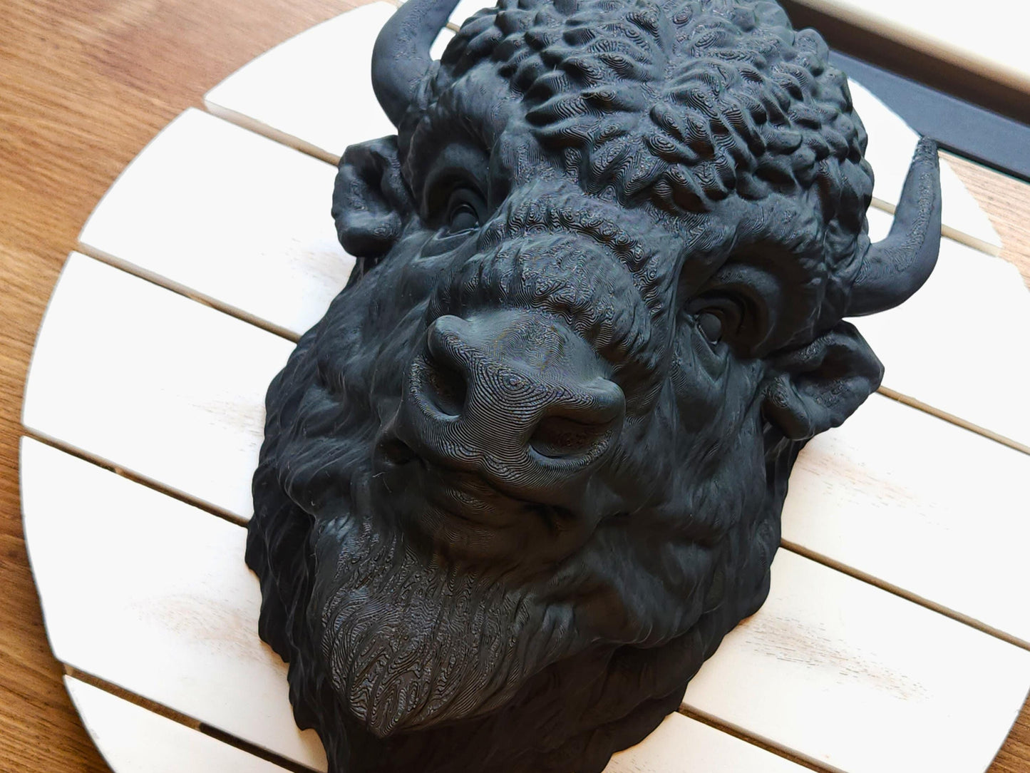 Three-quarter angle of a black buffalo head wall mount resting on a round white slatted board, showcasing its sculpted snout, curved horns, and textured fur details.