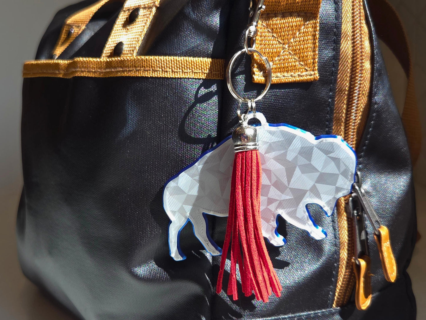 Buffalo football standing bison logo with geometric finish featured as a bag tag in silver with a blue backing, and a red tassel for contrast.