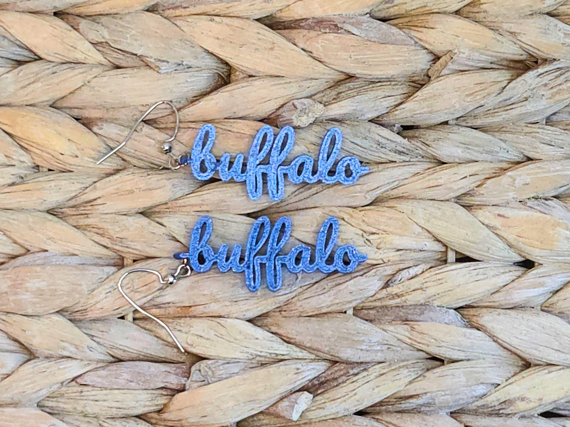 Pair of blue 3D printed script earrings spelling “buffalo” displayed on a woven background. Lightweight and perfect for Buffalo football fans.