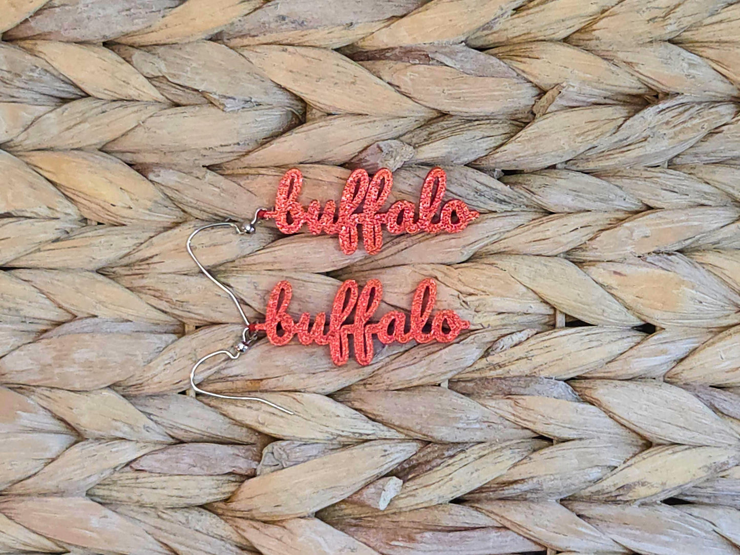 Pair of red 3D printed “buffalo” earrings with a glitter finish, styled on a neutral woven surface for a cozy game day look.