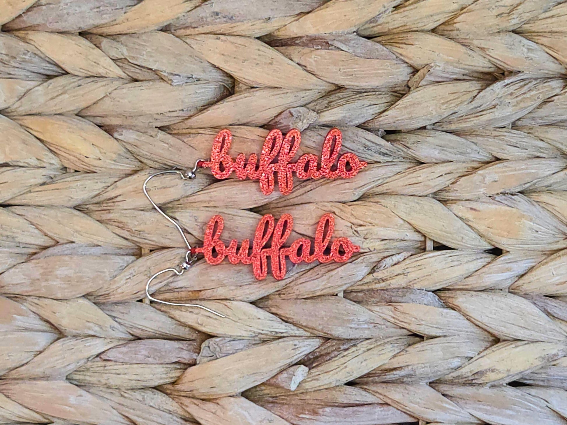 Pair of red 3D printed “buffalo” earrings with a glitter finish, styled on a neutral woven surface for a cozy game day look.