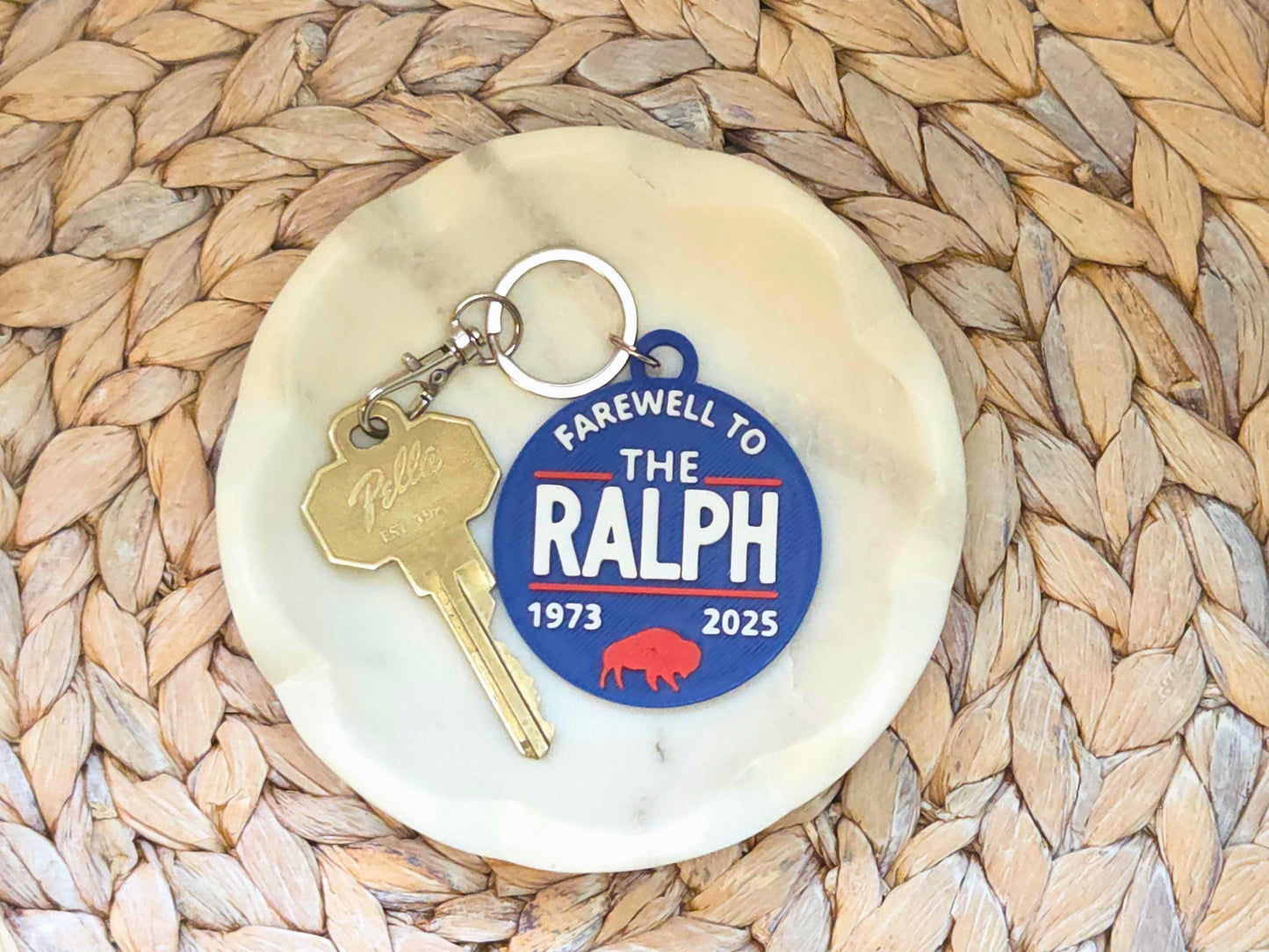 A top-down view of the commemorative keychain on a small white dish with a key attached. The keychain features bold white text, red accent lines, and a red buffalo icon, styled on a woven background.