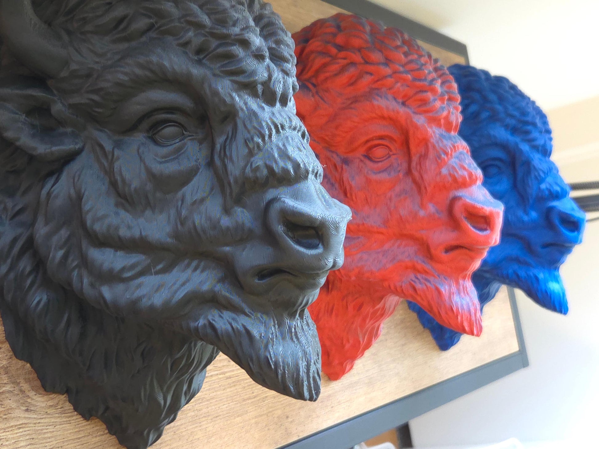 View of 3 3D printed bison heads in black, red/blue, and blue pictured side by side on a wooden background.