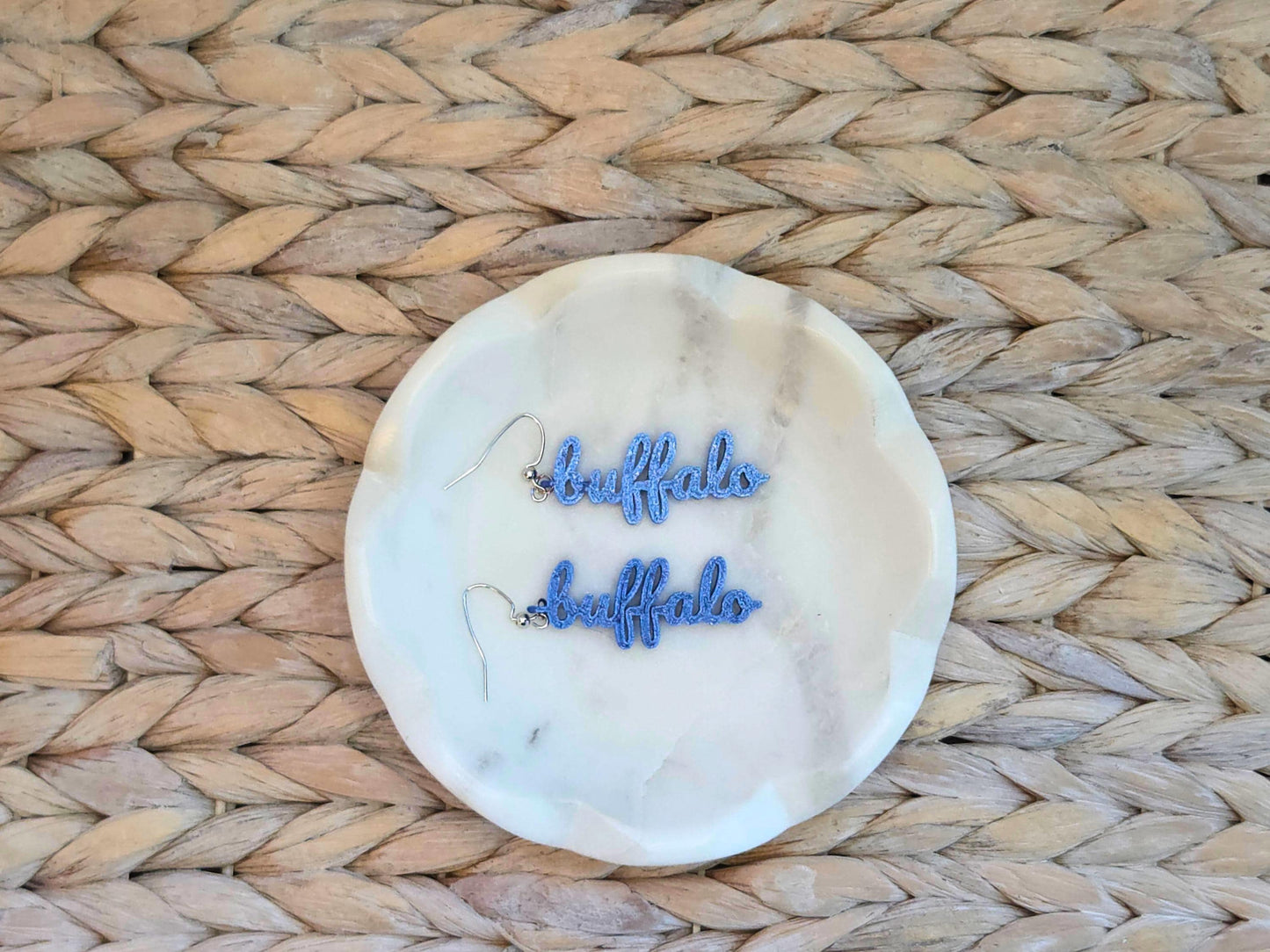 Blue “buffalo” script earrings placed on a white marble dish, highlighting their lightweight 3D printed texture and clean design.