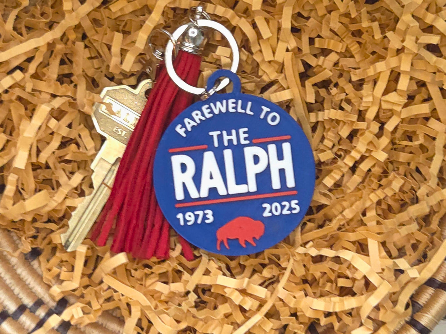 A close-up of a round, blue keychain with bold white text reading “Farewell to The Ralph 1973–2025,” featuring a red buffalo silhouette. The keychain is attached to a red tassel and brass key, resting on crinkled kraft paper