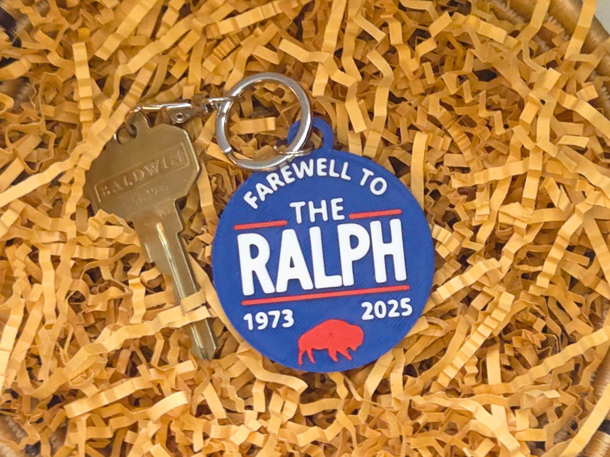 A keychain with “Farewell to The Ralph 1973–2025” design resting in crinkled kraft paper, attached to a brass key and keyring. Red buffalo graphic and raised white lettering are clearly visible