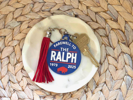 A blue “Farewell to The Ralph” keychain with red buffalo graphic and bold lettering, shown on a white dish with a red tassel and key. The dish sits on a woven mat for a clean, styled look.