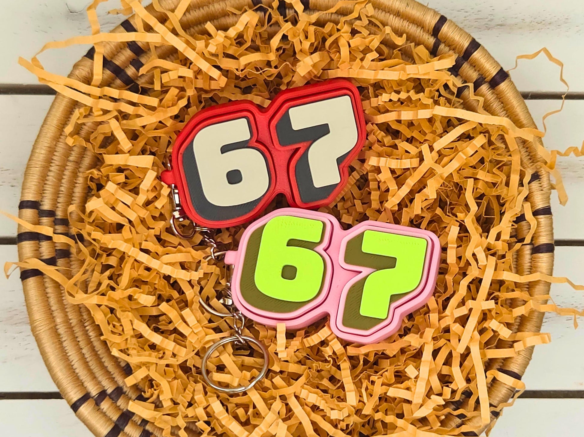 Picture of a red and tan and a pink and green fidget clicker keychain featuring the viral 67 meme positioned in some kraft paper on a wooden surface.