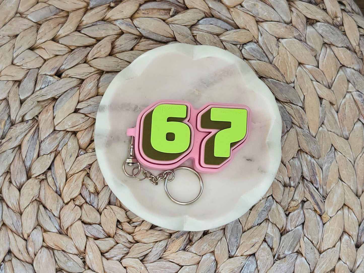 Picture of a pink and green fidget clicker keychain featuring the viral 67 meme in a dish on a woven surface.