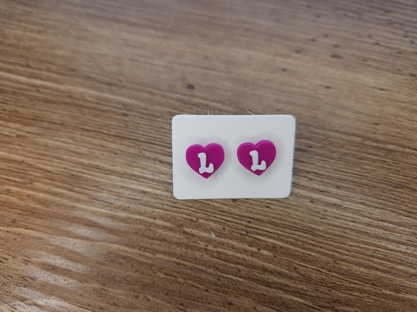 Close-up of magenta heart-shaped stud earrings with the letter &quot;L&quot; in white, displayed on a wooden surface.