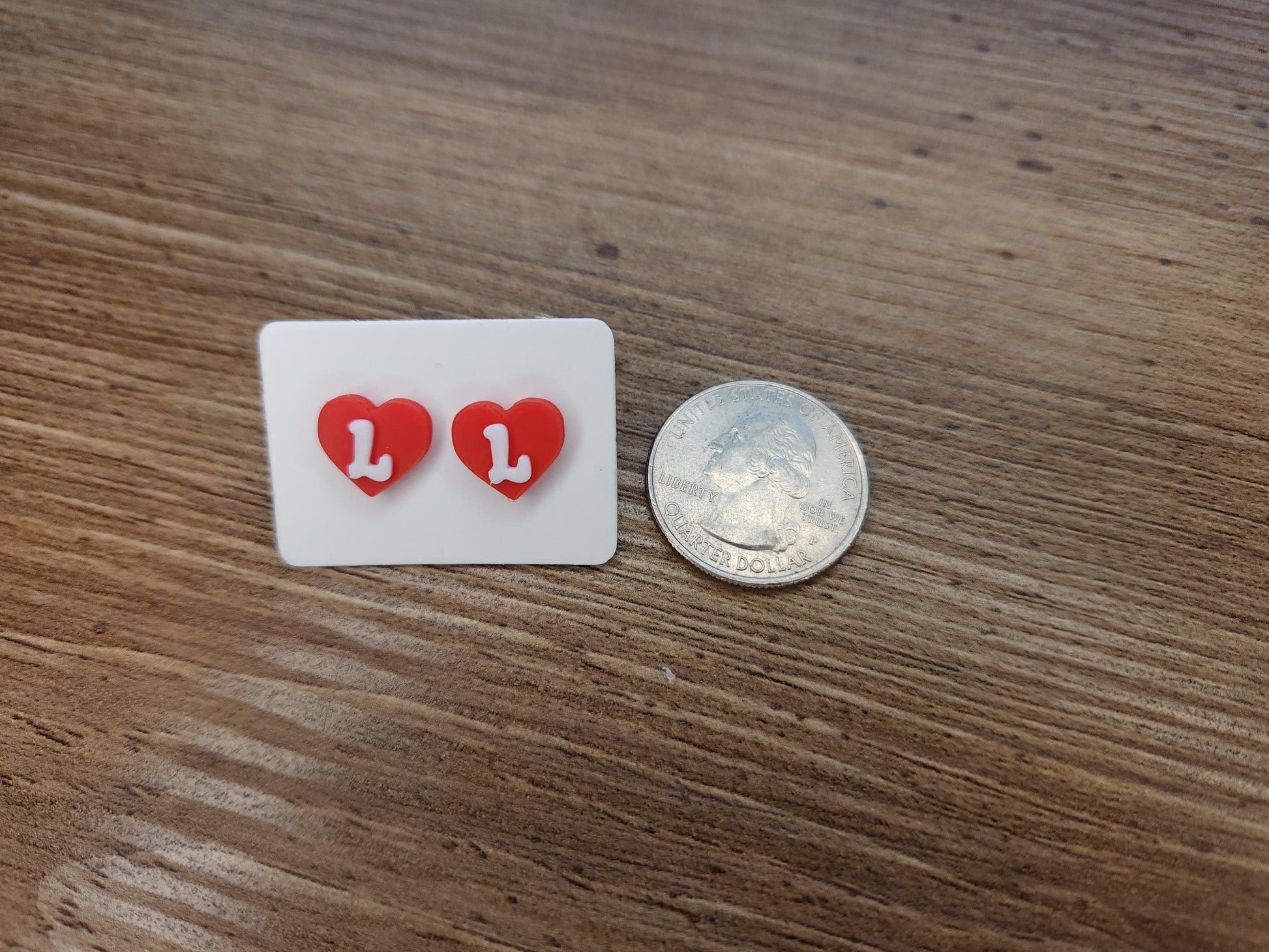 Red heart-shaped stud earrings with the letter &quot;L&quot; displayed on a white card next to a quarter for size reference, placed on a wooden surface.