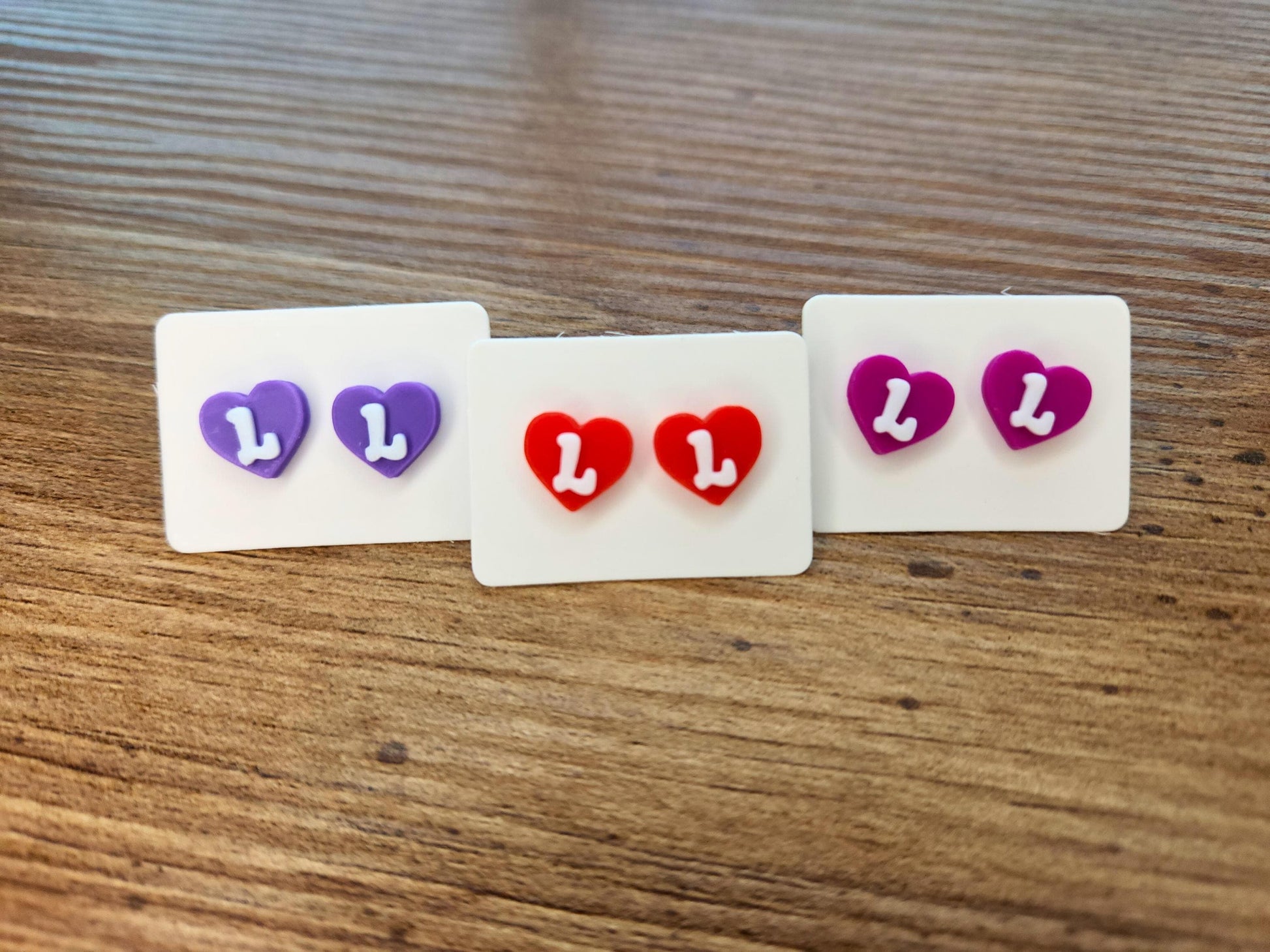 Three pairs of heart-shaped stud earrings in red, purple, and magenta, with the letter &quot;L&quot; in white, displayed on a wooden surface.