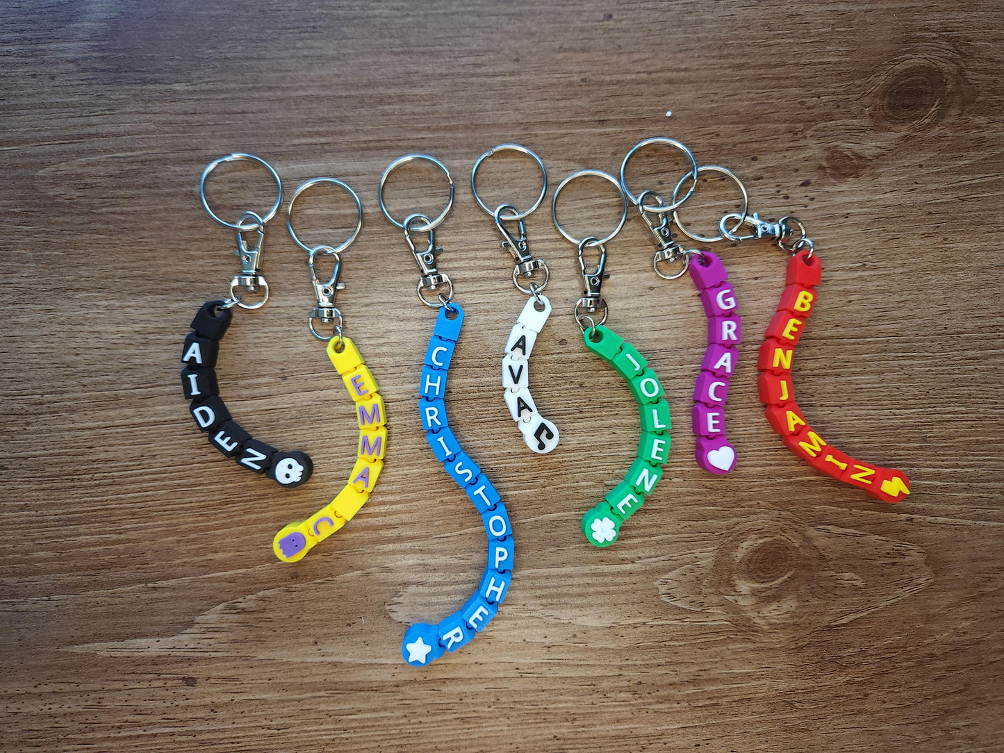 A collection of seven personalized keychains with names like &quot;AIDEN,&quot; &quot;EMMA,&quot; &quot;CHRISTOPHER,&quot; and more, each in different colors and adorned with unique symbols like stars and hearts. Ideal for backpacks, luggage, or gifts.