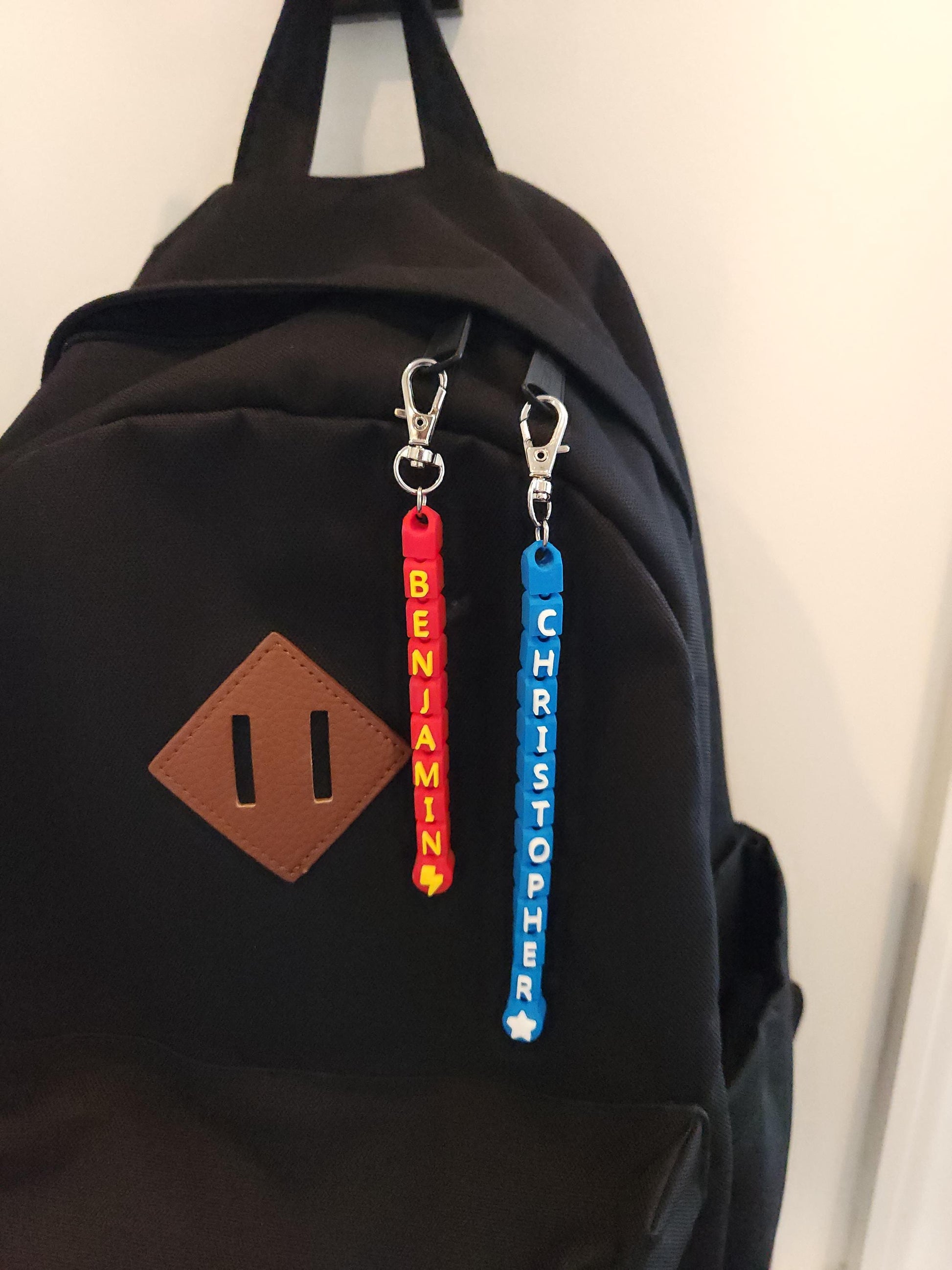 A black backpack featuring two vibrant personalized keychains—one in red and yellow spelling &quot;BENJAMIN&quot; with a lightning bolt and the other in blue spelling &quot;CHRISTOPHER&quot; with a star. Perfect for school or travel.