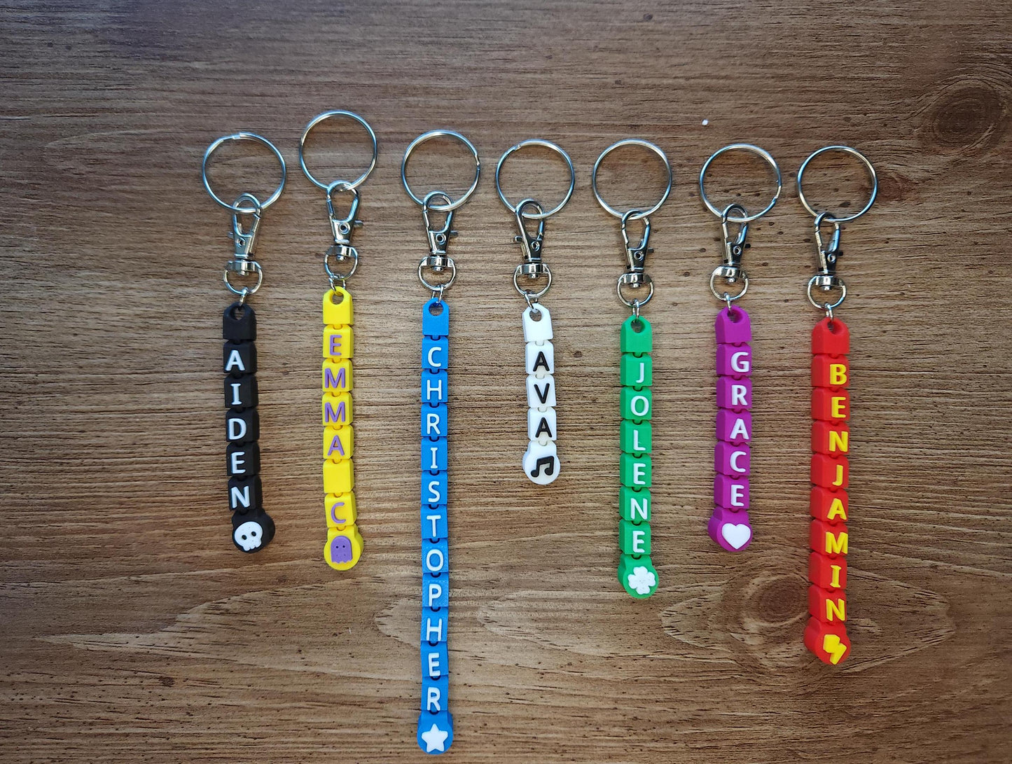 A set of seven straight keychains with names and vibrant colors, including black, yellow, blue, green, pink, and red. Each keychain is customizable, featuring symbols such as a skull, clover, or lightning bolt.