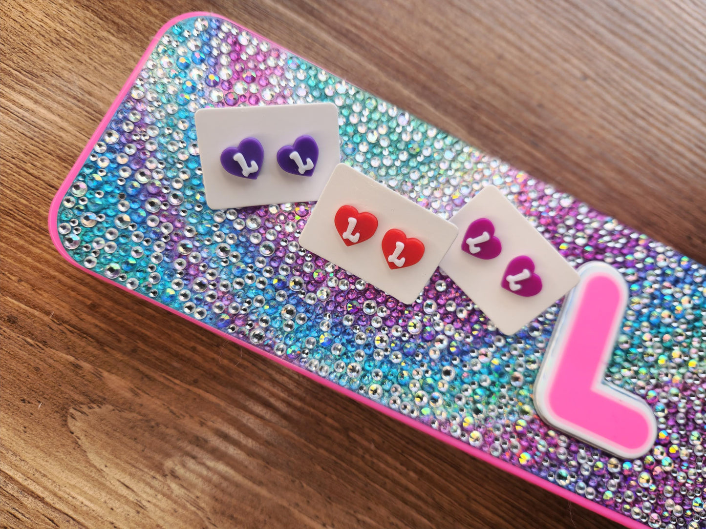 Three pairs of heart-shaped stud earrings in red, purple, and magenta with the letter &quot;L,&quot; displayed on a colorful, sequined background with a pink &quot;L&quot; decoration.