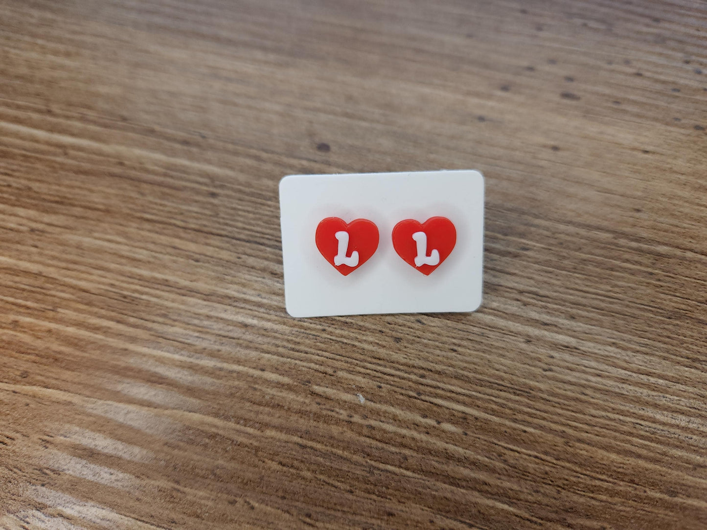 Close-up of red heart-shaped stud earrings with the letter &quot;L&quot; in white, displayed on a wooden surface.