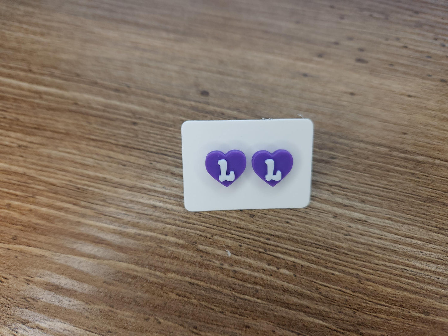 Close-up of purple heart-shaped stud earrings with the letter &quot;L&quot; in white, displayed on a wooden surface.